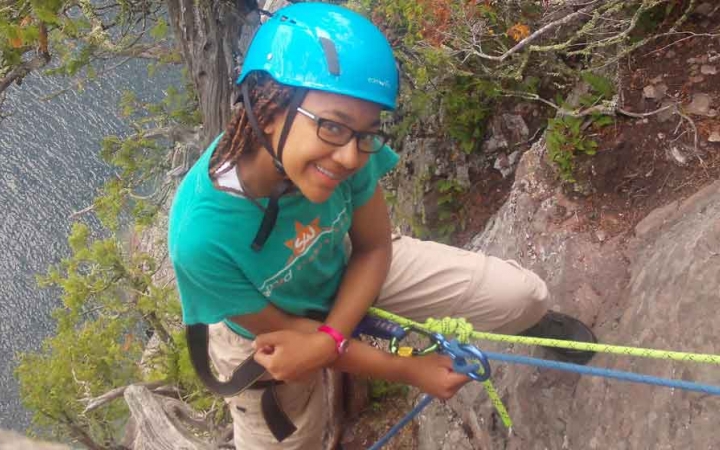 a student who is rock climbing above a body of water smiles at the camera